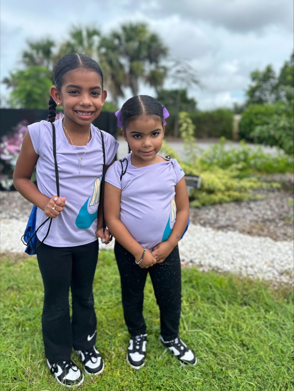 Two young girls in matching purple shirts and black pants pose together on the grass